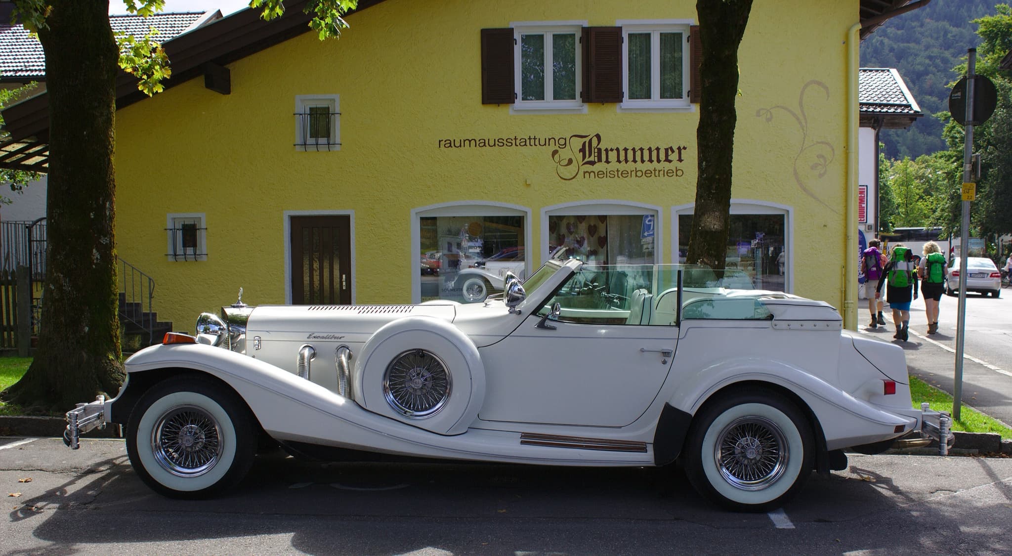 A white Excalibur phaeton with a side-mounted spare wheel and flowing wings, parked in a village street.