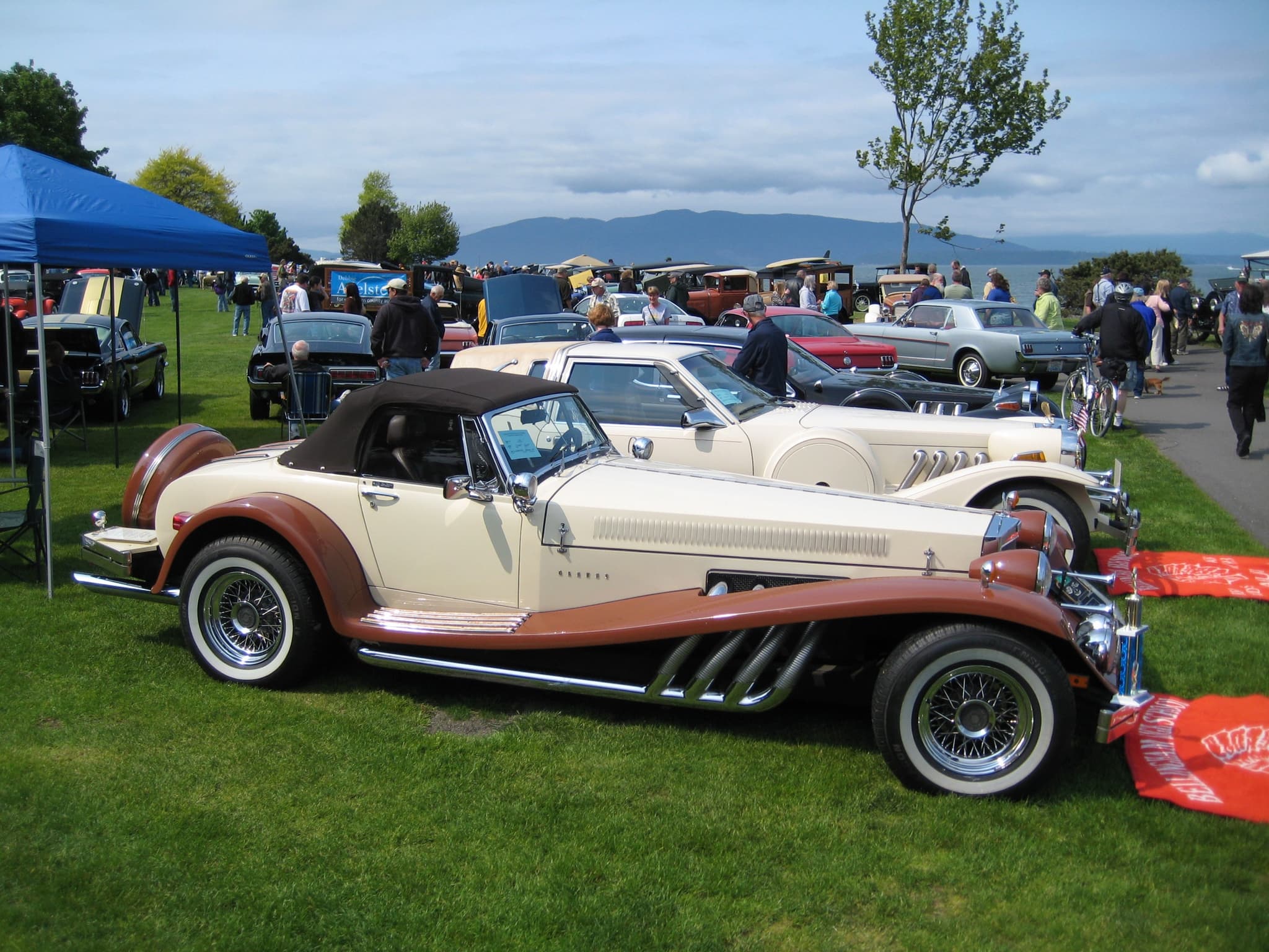A cream and brown Clénet roadster on a show lawn with a cream Golden Spirit coupé parked behind it.