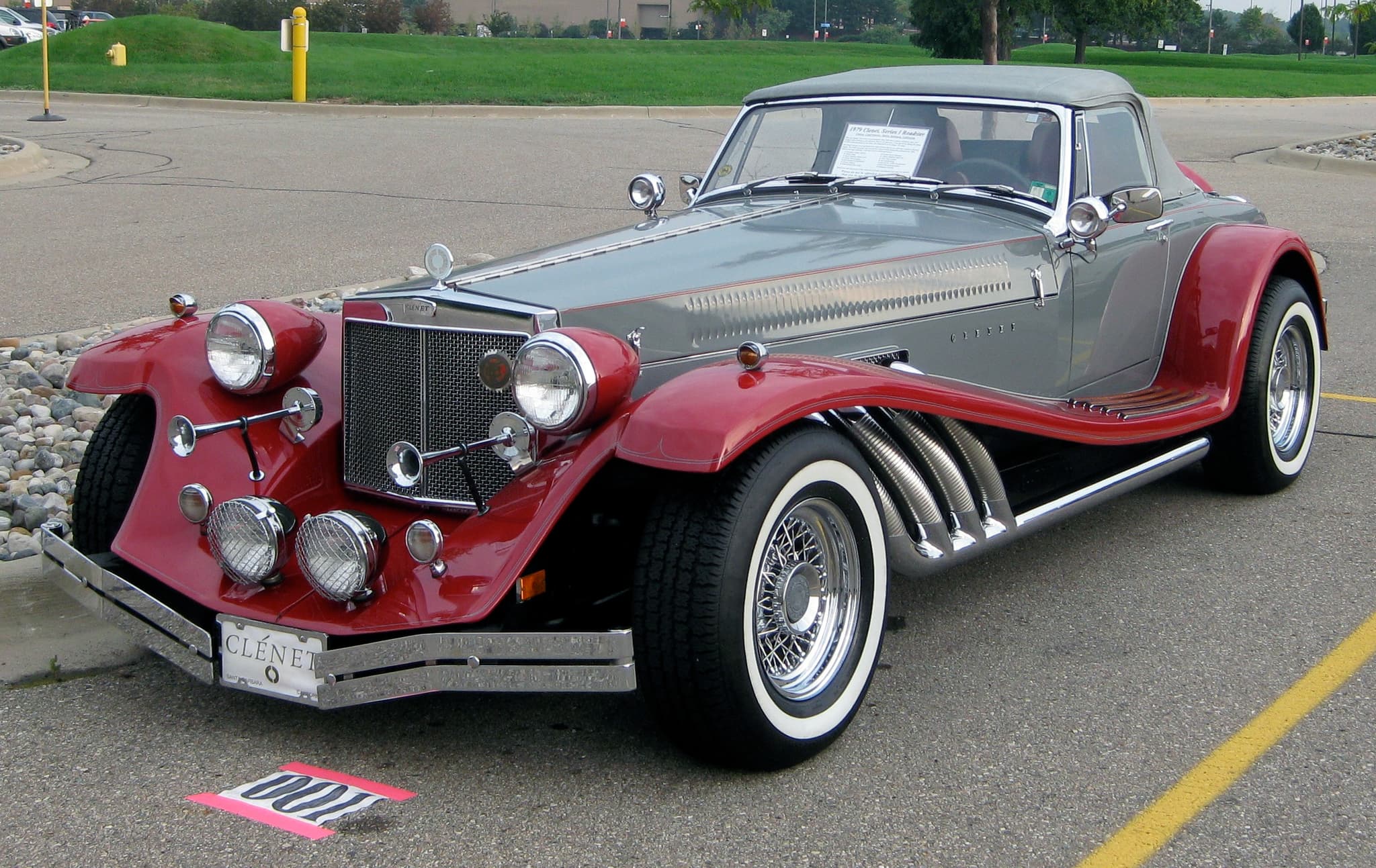 A grey and red Clénet roadster with flowing wings and chrome side pipes in a car park.