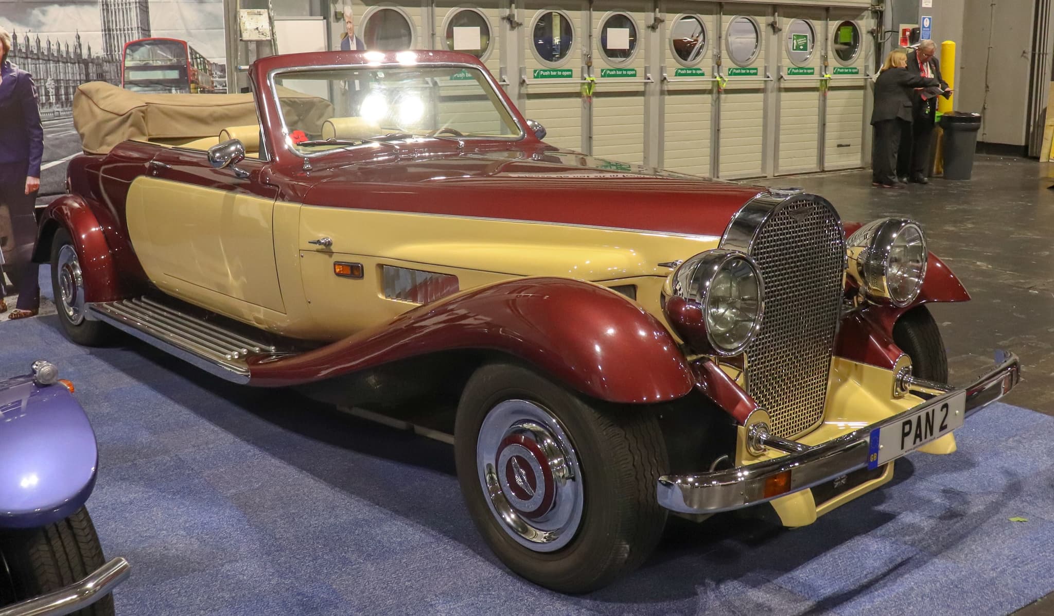 A maroon and cream Panther De Ville convertible with its hood folded, an upright mesh grille and separate headlamps, on a show stand.