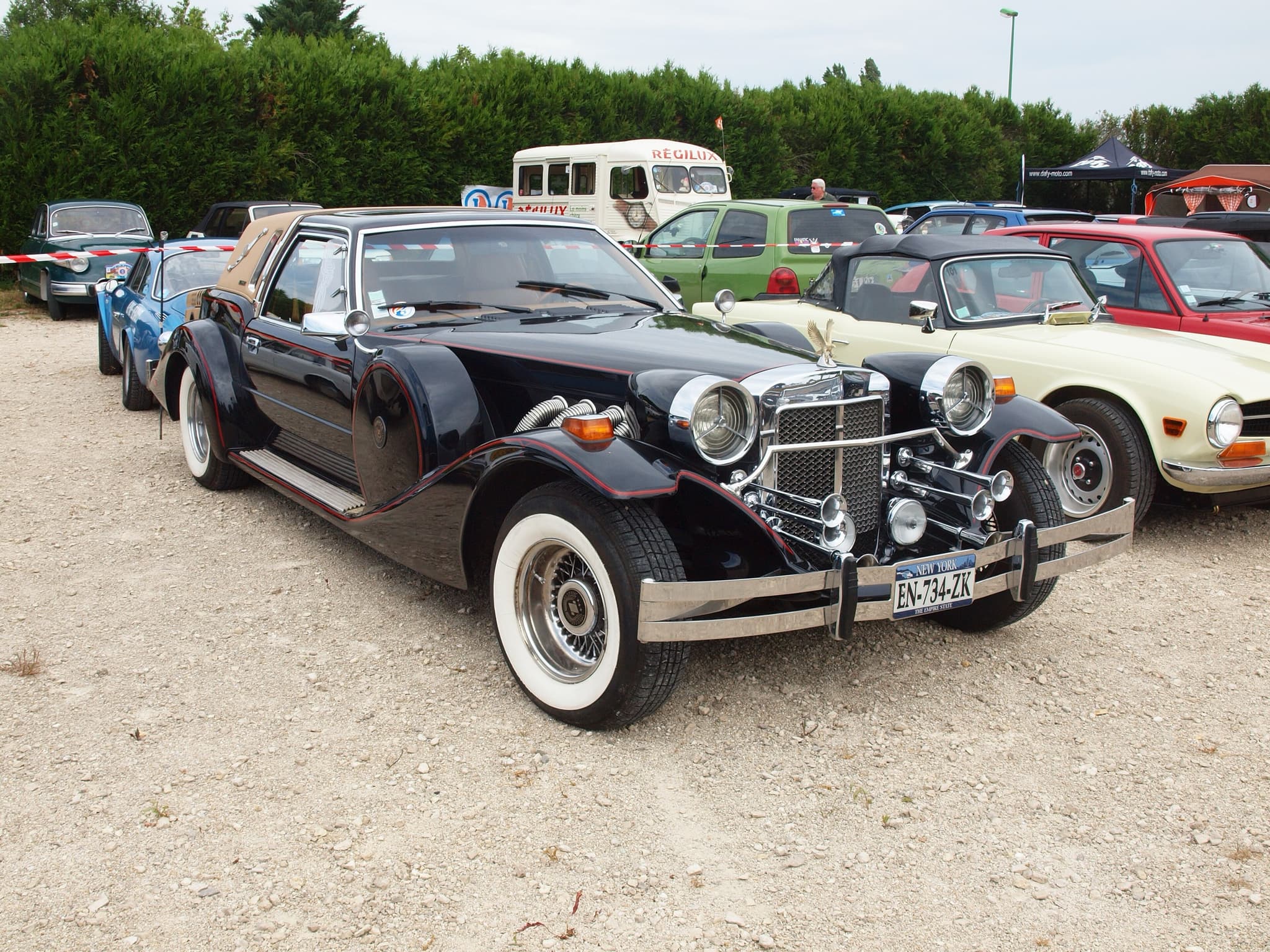 A black Golden Spirit coupé with red pinstripes, chrome trumpet horns and a tan padded rear roof, seen from the front three-quarter on a gravel show ground.